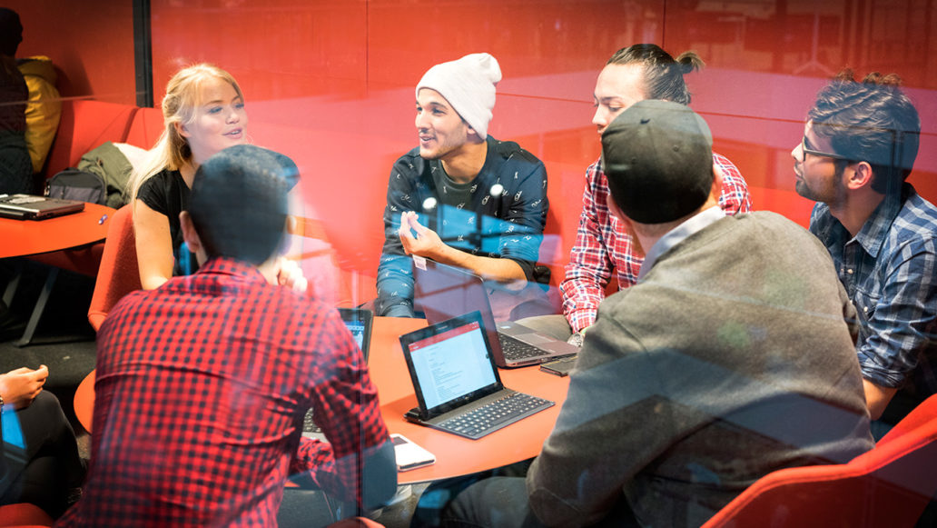 A group of students gathered around a table, discussing and studying on laptops. 