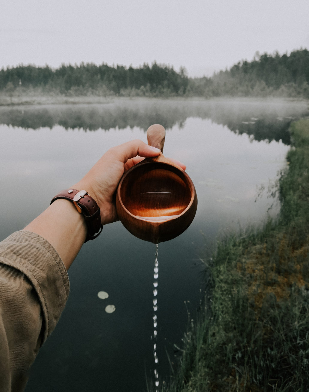 Woman pouring water from a kuksa wooden cup in front of a misty lake in the forest