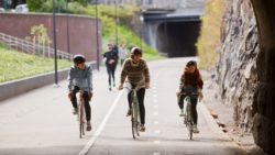 Three smiling people cycling on a wide street, people jogging behind them.