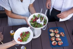 A person offering a bowl of salad for two people waiting, more food on the table.