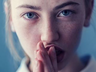 Close-up of a determined-looking person; she’s looking straight to the camera with her hands together in front of her mouth. 