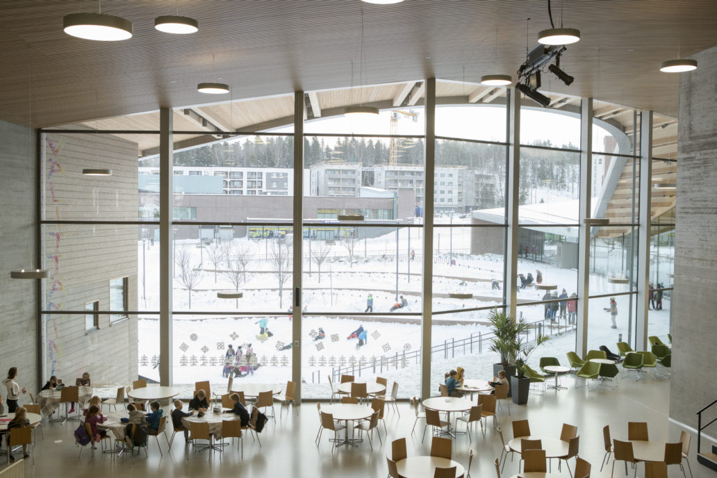 A school cafeteria has some pupils eating lunch in front of a big window. 