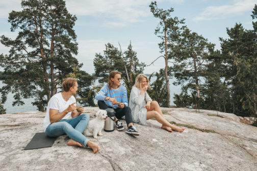 Three young adults, two men and a woman, with a white poodle sitting on smooth bedrock drinking coffee out of guksis and smiling with pine trees and a summery lake in the background