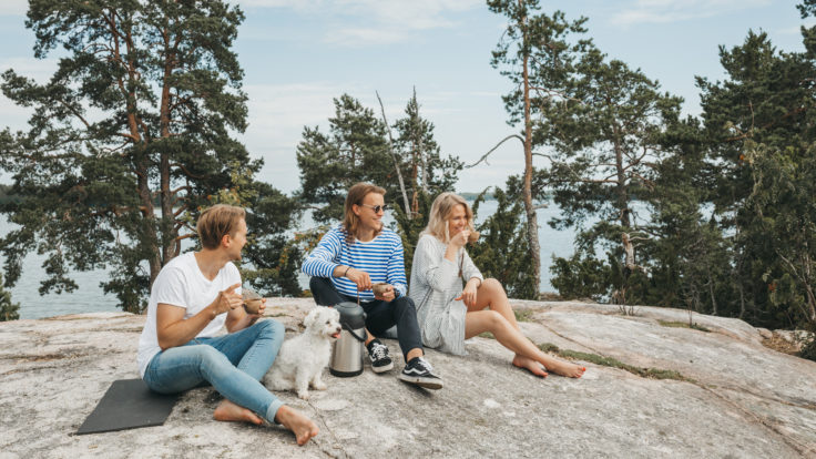 Three young adults, two men and a woman, with a white poodle sitting on smooth bedrock drinking coffee out of guksis and smiling with pine trees and a summery lake in the background