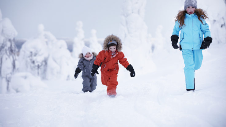Three children of different ages running in thick winter wear in a snow-covered landscape