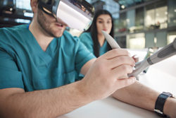 A man in green scrubs and VR glasses operating medical equipment with woman watching in background
