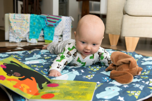 A baby on tummy time looking at a book with a maternity package and baby clothes in the background