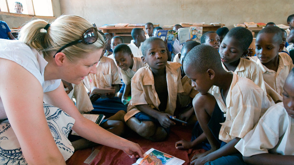 A classroom filled with young children sitting on the floor and a teacher pointing out something from a book they’re reading. 