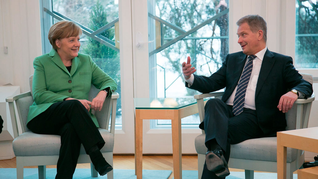 Angela Merkel and Sauli Niinistö sitting in armchairs in front of big windows and discussing. 