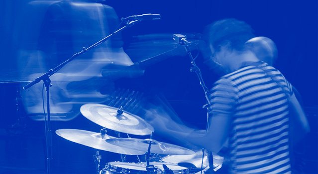 A blurred photo of a drummer drumming their kit and a guitarist playing in the background. 