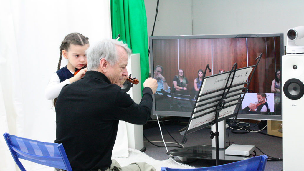 An elderly man teaching a young girl how to play the violin, online students watching them are visible on a big screen. 