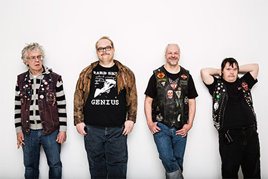 Four punk rocker men dressed in jeans and vests leaning on a white wall. 