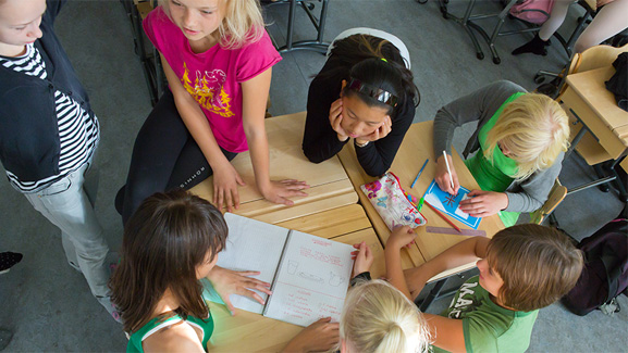 Children in a classroom, gathered around their desks studying.