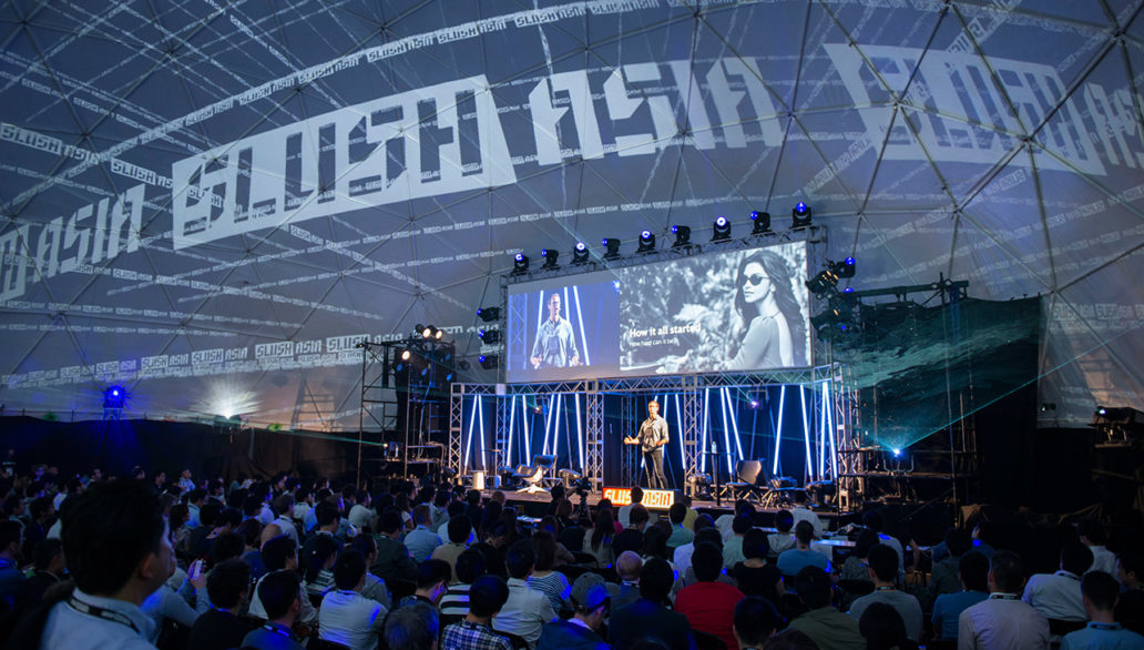 A photo from Slush Japan event shows a large crowd of people gathered by a stage where someone is presenting. 