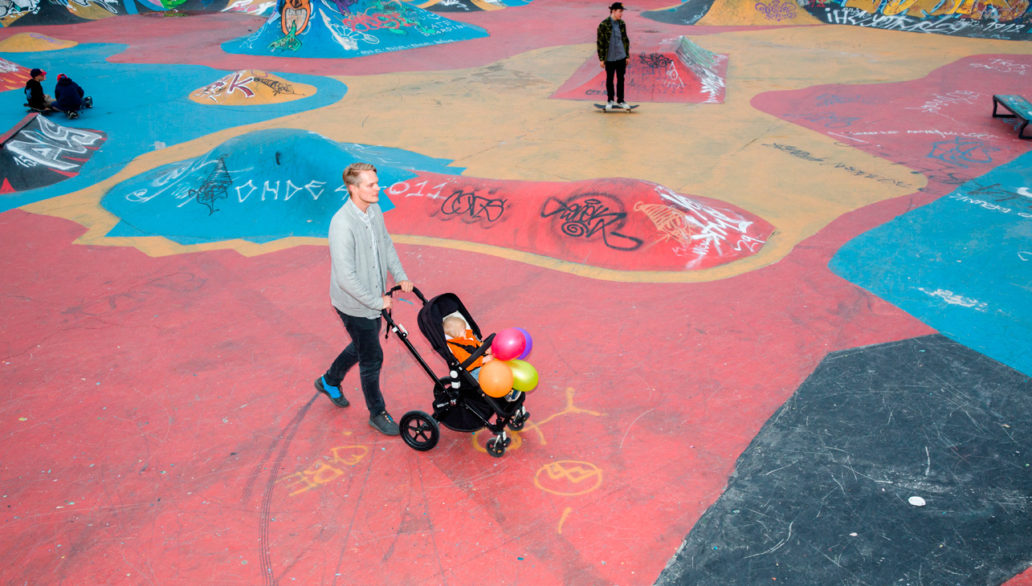 A man pushing a stroller at a colourful skate park with skaters in the background. 