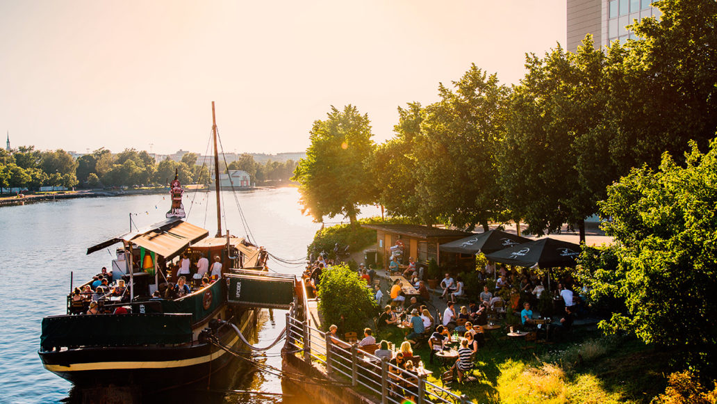 A docked restaurant boat filled with people, more people are sitting at tables on the shore. 